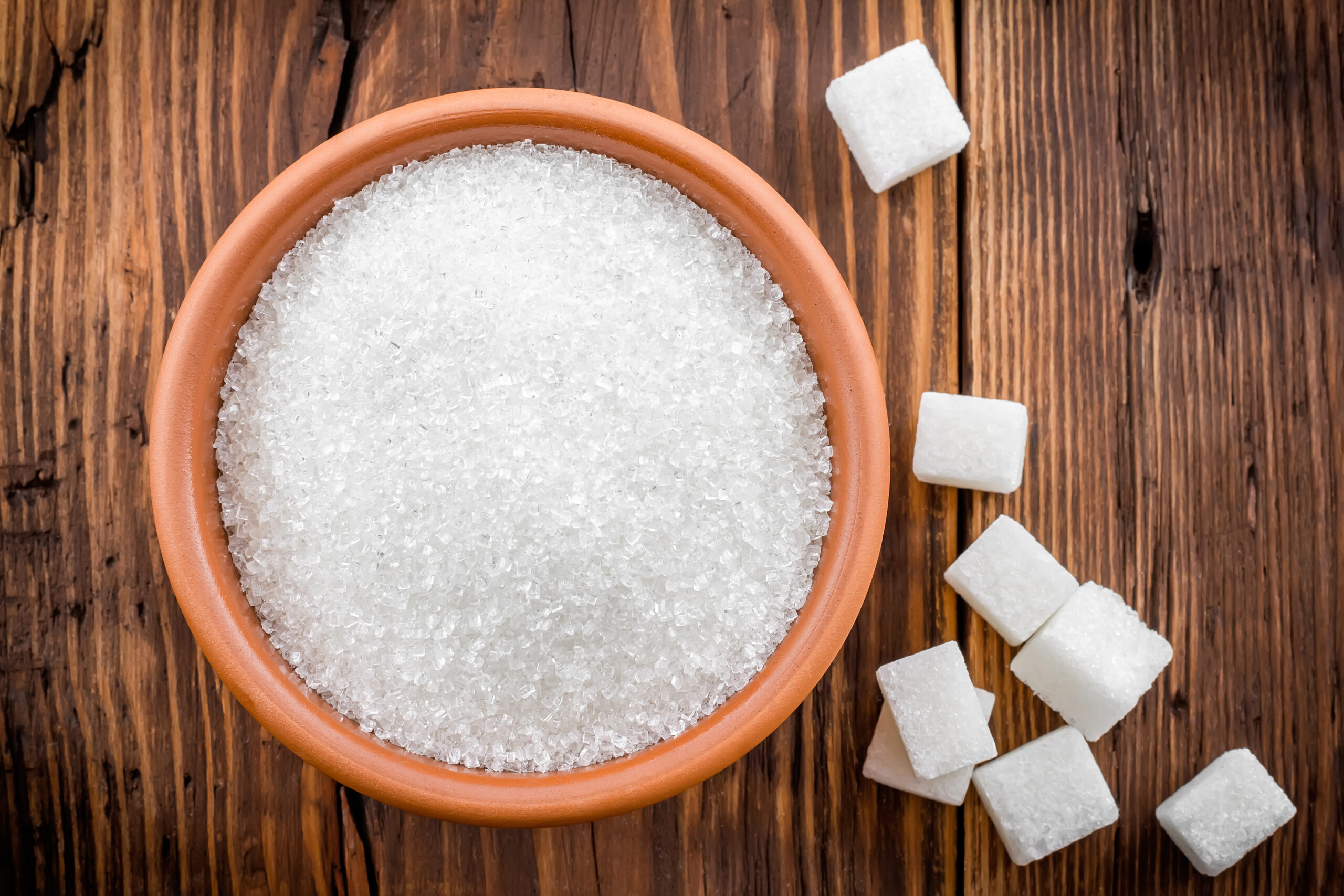 Sugar A terracotta bowl filled with coarse white sugar granules on a rustic wooden table, surrounded by scattered sugar cubes.